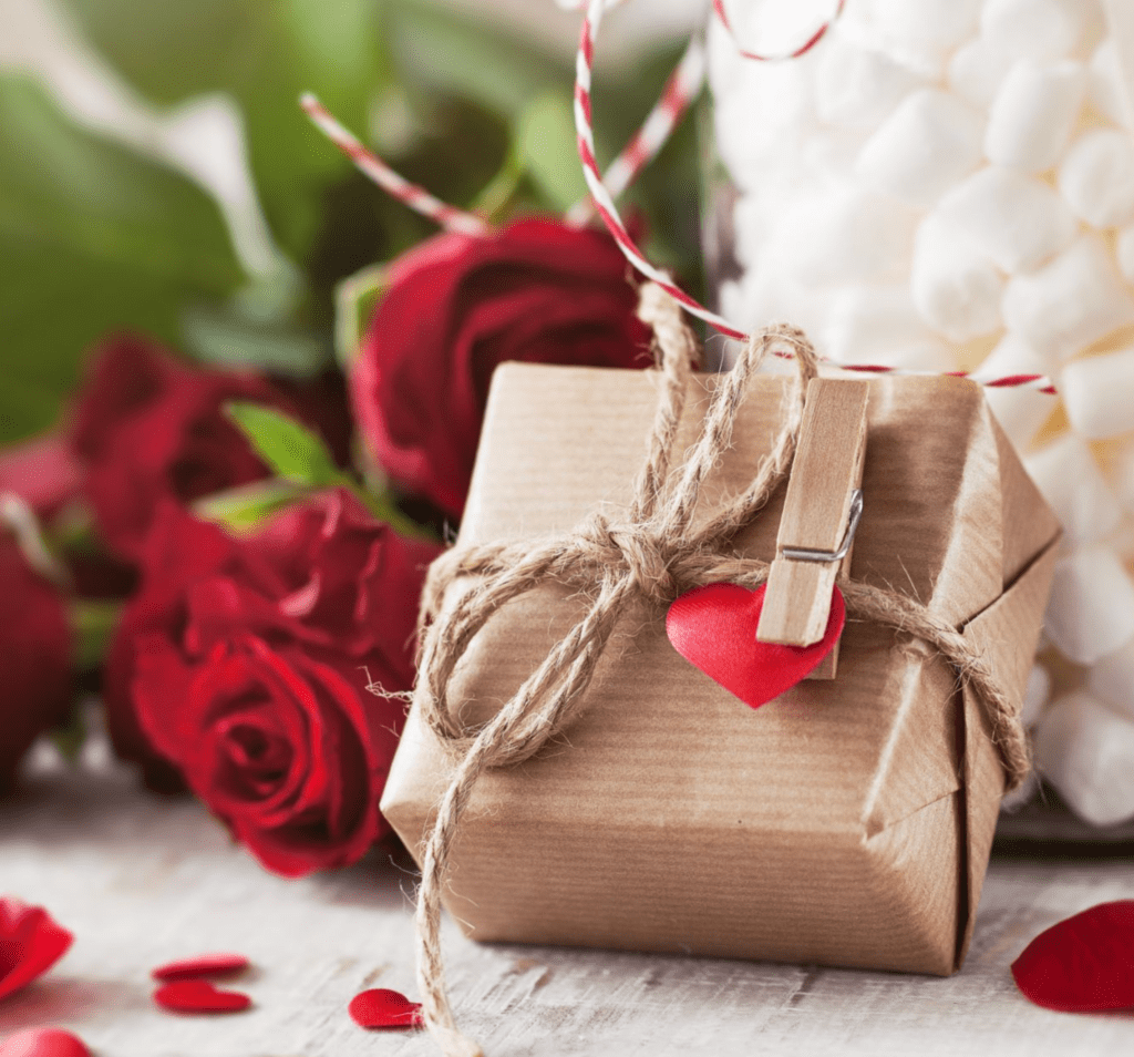 A imagem mostra um presente embrulhado em papel kraft, amarrado com barbante rústico e decorado com um pequeno coração vermelho preso por um mini pregador de madeira. Ao fundo, há rosas vermelhas e marshmallows brancos, compondo um cenário romântico, típico de celebrações como o Dia dos Namorados. Pétalas e elementos decorativos completam a composição sobre uma superfície de madeira clara.
